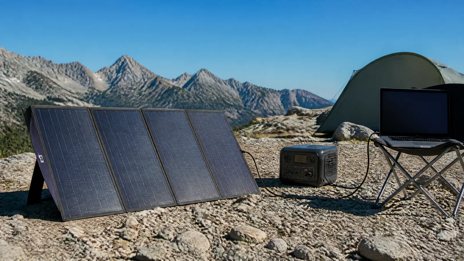 Foldable portable solar panel angled toward the sun on rocky ground, charging a portable power station and laptop at an off-grid mountain campsite